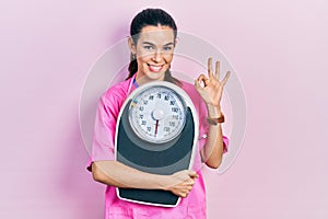 Young brunette nutritionist woman holding scale doing ok sign with fingers, smiling friendly gesturing excellent symbol