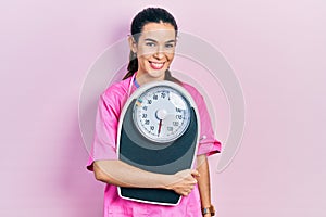 Young brunette nutritionist woman holding scale looking positive and happy standing and smiling with a confident smile showing