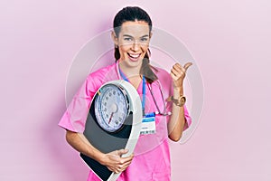 Young brunette nutritionist woman holding scale pointing thumb up to the side smiling happy with open mouth
