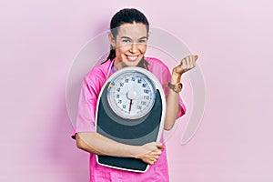 Young brunette nutritionist woman holding scale screaming proud, celebrating victory and success very excited with raised arm