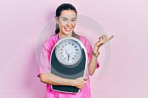 Young brunette nutritionist woman holding scale smiling happy pointing with hand and finger to the side