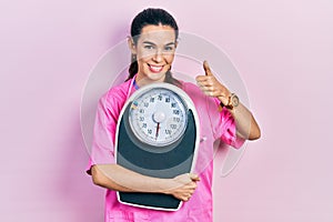 Young brunette nutritionist woman holding scale smiling happy and positive, thumb up doing excellent and approval sign