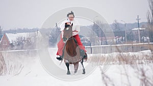 Young brunette in a red dress galloping fast on a horse through the snow-covered field in the winter