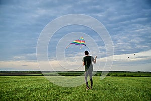 Young brunette skinny man, wearing dark green t-shirt, playing with colorful kite on green field meadow in summer. Kite flying in