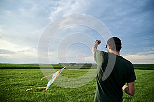 Young brunette skinny man, wearing dark green t-shirt, playing with colorful kite on green field meadow in summer. Kite flying in