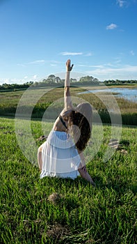 Young brunette spanish woman doing reverse warrior yoga pose in a field next to a lake with long curly hair