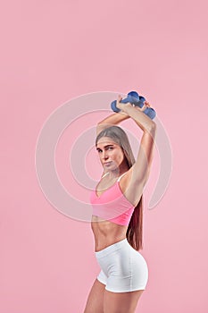 Young brunette sporty woman posing or training with dumm bell in hands, portrait, sport outfit on pink background.