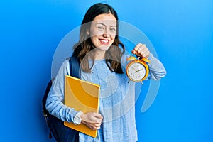 Young brunette student woman holding alarm clock smiling with a happy and cool smile on face