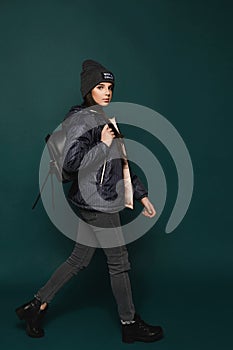 Young brunette teen girl, in jacket and hat with a bag, walking over dark-green background,