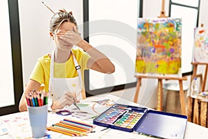 Young brunette teenager at art studio covering eyes with hand, looking serious and sad