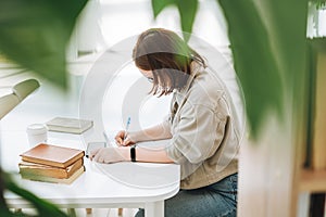 Young brunette teenager girl college student in glasses doing homework with mobile phone at modern library public place, green