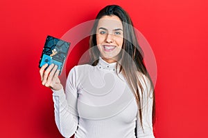 Young brunette teenager holding hdd disk looking positive and happy standing and smiling with a confident smile showing teeth