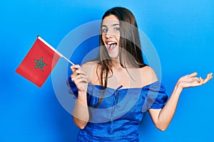 Young brunette teenager holding morocco flag celebrating achievement with happy smile and winner expression with raised hand