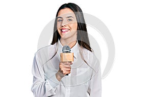 Young brunette teenager holding reporter microphone looking positive and happy standing and smiling with a confident smile showing