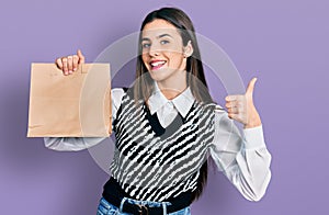 Young brunette teenager holding take away paper bag smiling happy and positive, thumb up doing excellent and approval sign