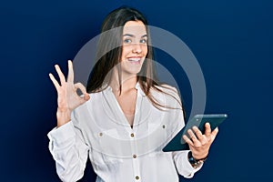 Young brunette teenager using touchpad device doing ok sign with fingers, smiling friendly gesturing excellent symbol