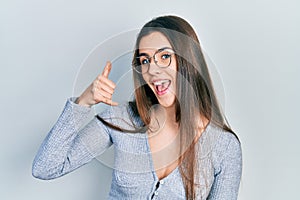 Young brunette teenager wearing casual sweater and glasses smiling doing phone gesture with hand and fingers like talking on the