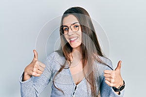 Young brunette teenager wearing casual sweater and glasses success sign doing positive gesture with hand, thumbs up smiling and