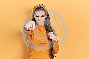 Young brunette teenager wearing casual yellow sweater punching fist to fight, aggressive and angry attack, threat and violence