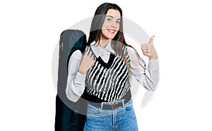 Young brunette teenager wearing guitar case smiling happy and positive, thumb up doing excellent and approval sign