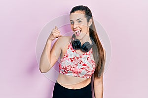 Young brunette teenager wearing gym clothes and using headphones smiling doing phone gesture with hand and fingers like talking on