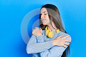 Young brunette teenager wearing headphones on neck hugging oneself happy and positive, smiling confident