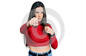 Young brunette teenager wearing red turtleneck sweater punching fist to fight, aggressive and angry attack, threat and violence