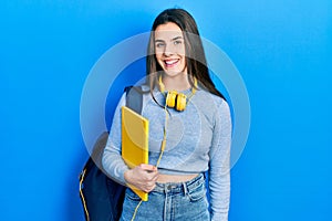 Young brunette teenager wearing student backpack and headphones looking positive and happy standing and smiling with a confident