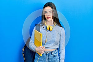 Young brunette teenager wearing student backpack and headphones thinking attitude and sober expression looking self confident