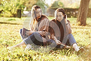 Young brunette twin sisters sitting close to each other with eyes closed on the grass,legs slightly bent in knees and