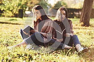 Young brunette twin sisters sitting close to each other on the grass with legs slightly bent in knees and crossed