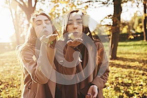 Young brunette twin sisters standing close to each other and blowing confetti into camera, wearing casual coat in autumn