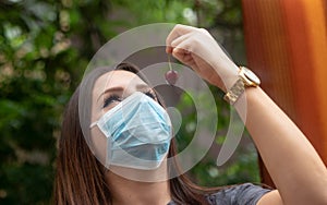 Young brunette wearing a surgical face mask holding a red cherry trying to eat it. Gesturing towards it outside on green