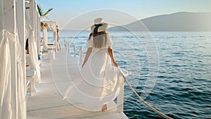 Young brunette in white dress, hat, and flowing hair runs on wooden pier by sunset at sea. Evokes tourism, summertime