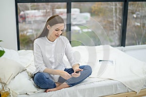 A young brunette in a white shirt and jeans writes SMS on the phone. A girl sits on the bed and reads the message text