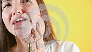 Young brunette who checks her skin with magnifying glass on yellow background. Skin allergy concept, facial rashes