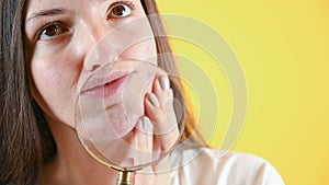 Young brunette who checks her skin on yellow background. Woman with problem skin holding magnifying glass by face.