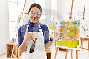 Young brunette woman at art studio showing palm hand and doing ok gesture with thumbs up, smiling happy and cheerful