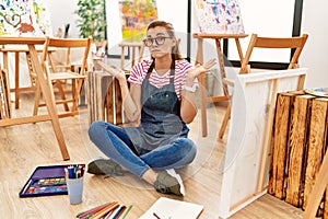 Young brunette woman at art studio sitting on the floor clueless and confused expression with arms and hands raised