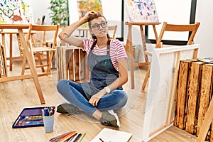 Young brunette woman at art studio sitting on the floor confuse and wonder about question