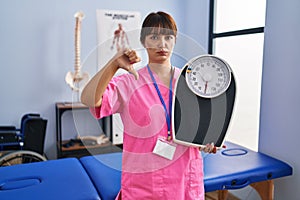 Young brunette woman as nutritionist holding weighing machine with angry face, negative sign showing dislike with thumbs down,
