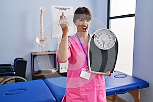 Young brunette woman as nutritionist holding weighing machine annoyed and frustrated shouting with anger, yelling crazy with anger