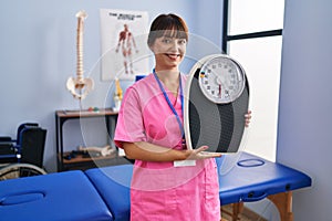 Young brunette woman as nutritionist holding weighing machine looking positive and happy standing and smiling with a confident