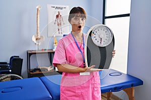 Young brunette woman as nutritionist holding weighing machine scared and amazed with open mouth for surprise, disbelief face