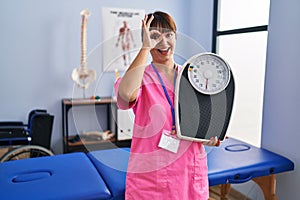 Young brunette woman as nutritionist holding weighing machine smiling happy doing ok sign with hand on eye looking through fingers