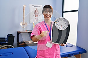 Young brunette woman as nutritionist holding weighing machine smiling happy pointing with hand and finger