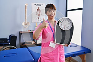 Young brunette woman as nutritionist holding weighing machine smiling happy and positive, thumb up doing excellent and approval