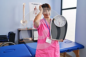 Young brunette woman as nutritionist holding weighing machine stressed and frustrated with hand on head, surprised and angry face