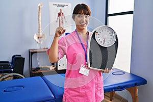 Young brunette woman as nutritionist holding weighing machine surprised with an idea or question pointing finger with happy face,