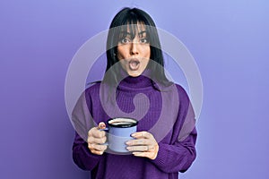 Young brunette woman with bangs drinking a cup of coffee scared and amazed with open mouth for surprise, disbelief face